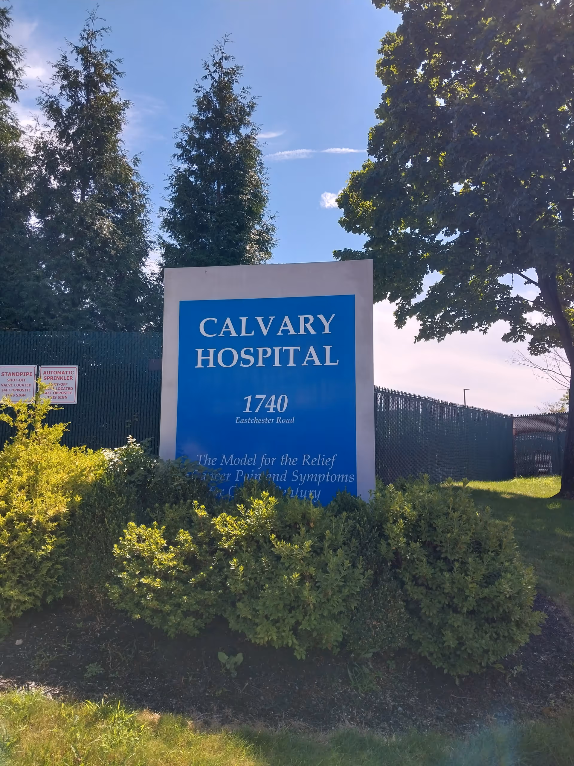 A blue Calvary Hospital sign reading 1740 Eastchester Road stands among shrubs.