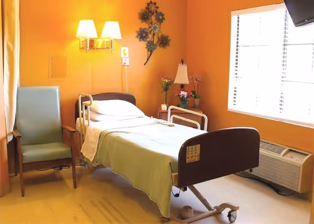 A hospital-style bed in a warmly painted private room with a recliner, bedside table with lamp and flowers, and a window with blinds.