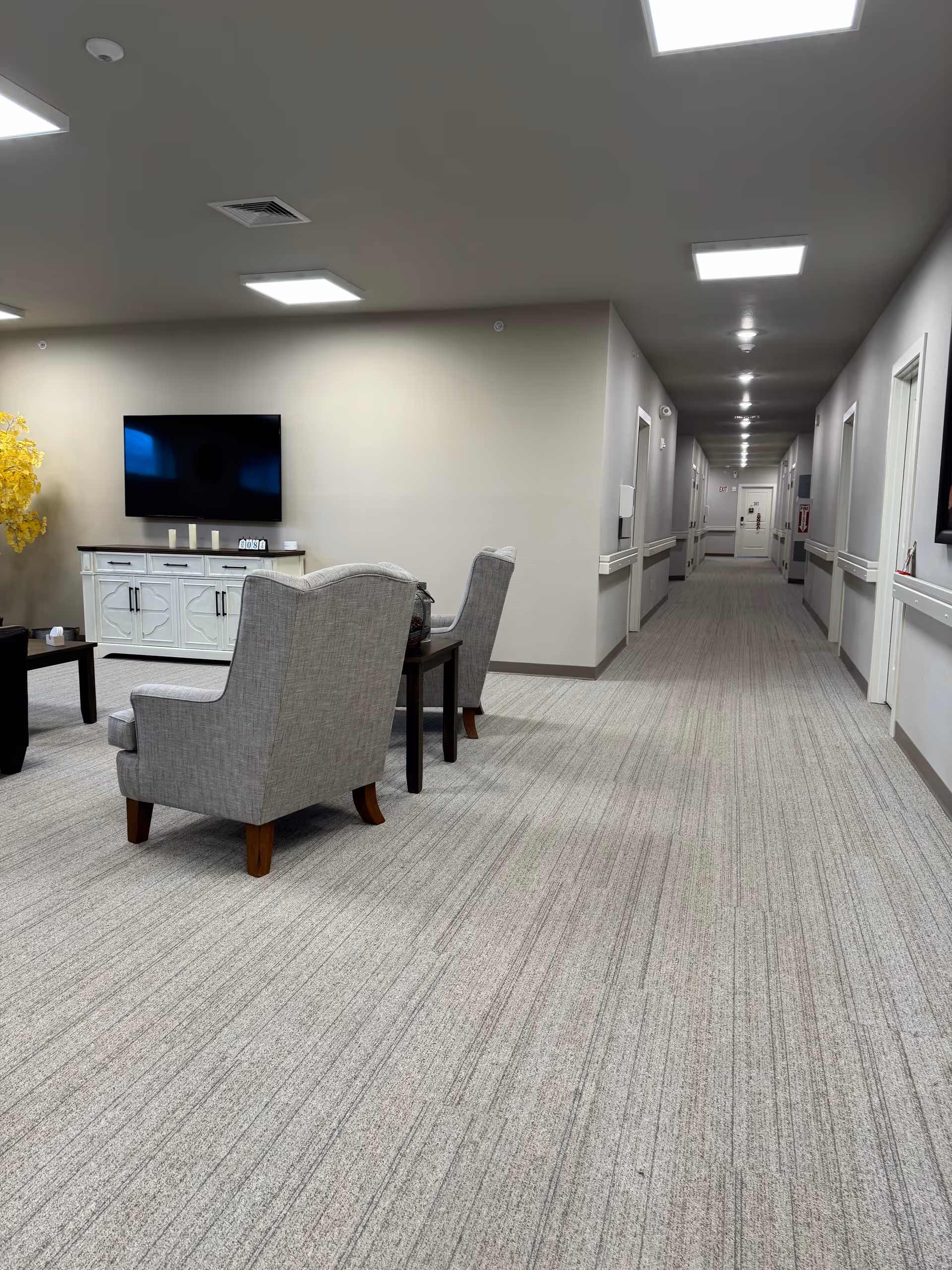 Lobby lounge with two gray armchairs, a TV, and a long forward hallway.