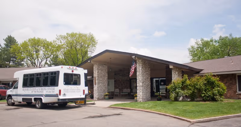 Exterior view of the Rehabilitation and Nursing Center of the Rockies building with a covered entrance supported by stone pillars. A white shuttle bus with the facility's name is parked in front. There is a well-maintained lawn and some bushes near the entrance, and an American flag is hanging from the covered area.