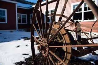 Close-up view of a large rusty metal wheel, possibly part of old farming equipment, situated outdoors on a patch of snow-covered ground with a red building and windows in the background.