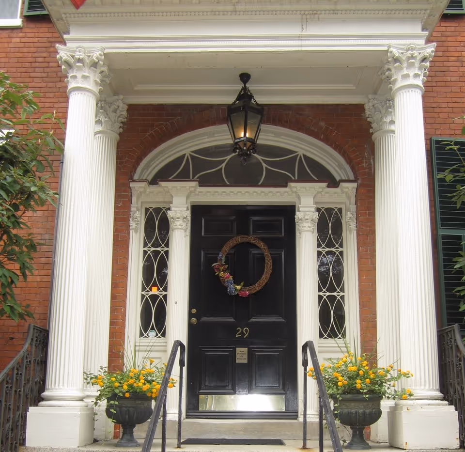 Front entrance of a brick building with a black door numbered 29, flanked by white decorative columns and sidelights with intricate window designs. A wreath with flowers hangs on the door, and two planters with yellow flowers are placed symmetrically on either side of the steps leading up to the door. A black lantern hangs above the door.