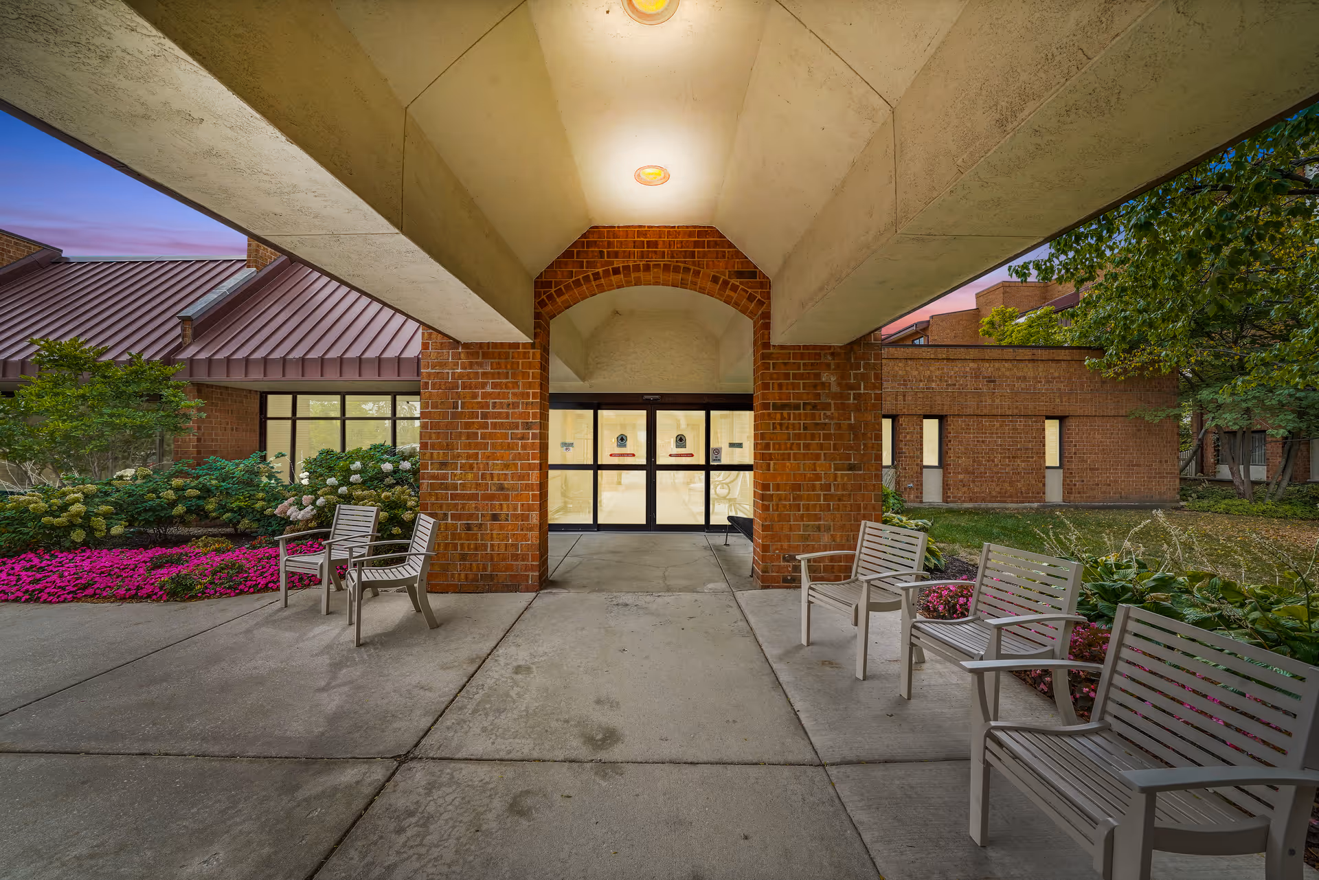 Covered entrance and walkway to a brick building with outdoor chairs and landscaped flower beds.