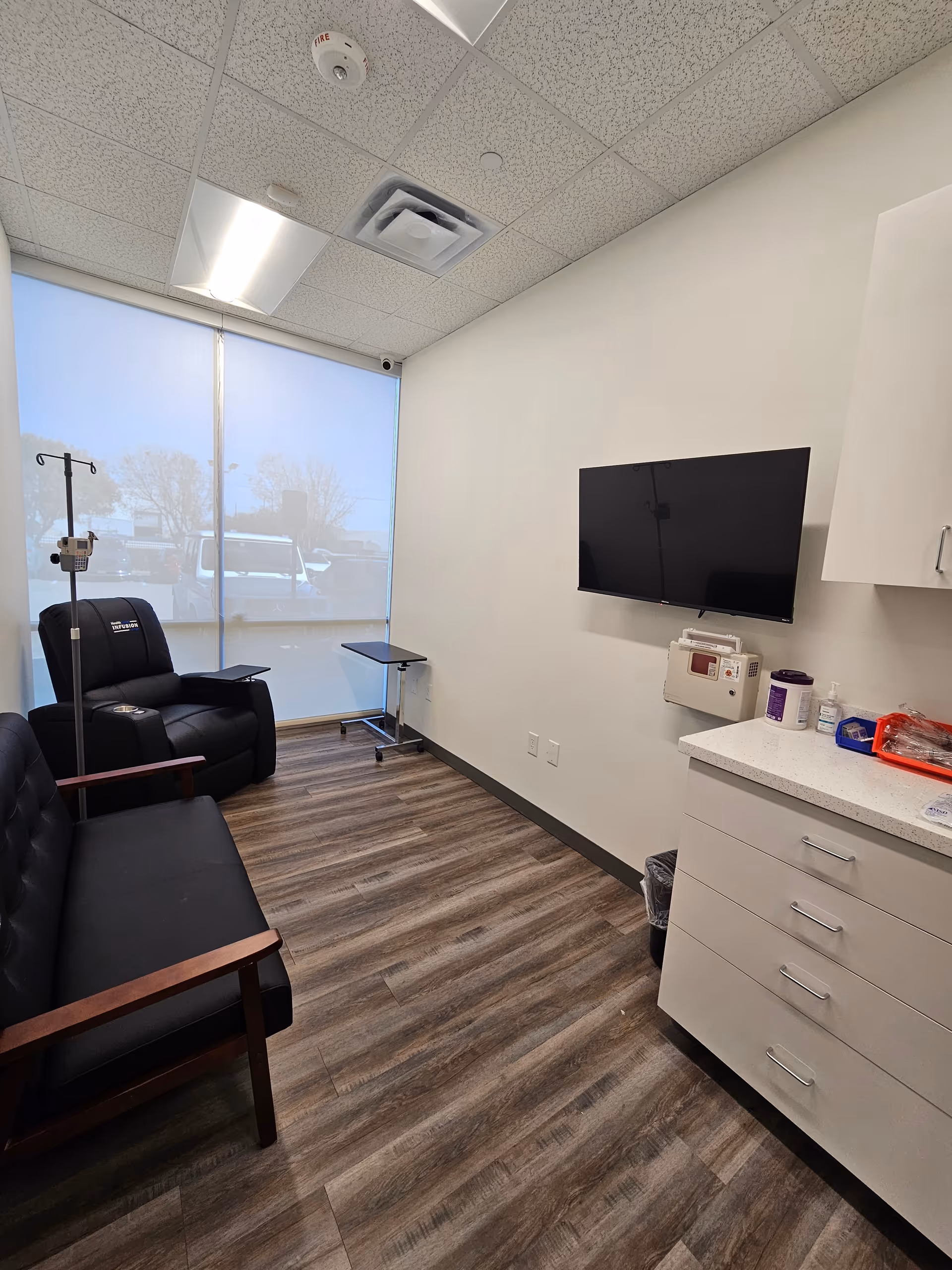 A clinical infusion room with black leather chairs, IV stand, TV, and medical supplies.