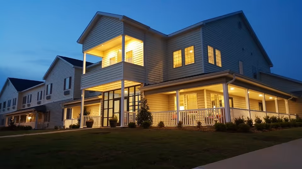 Exterior view of a two-story senior living facility with a lit wraparound porch at dusk.