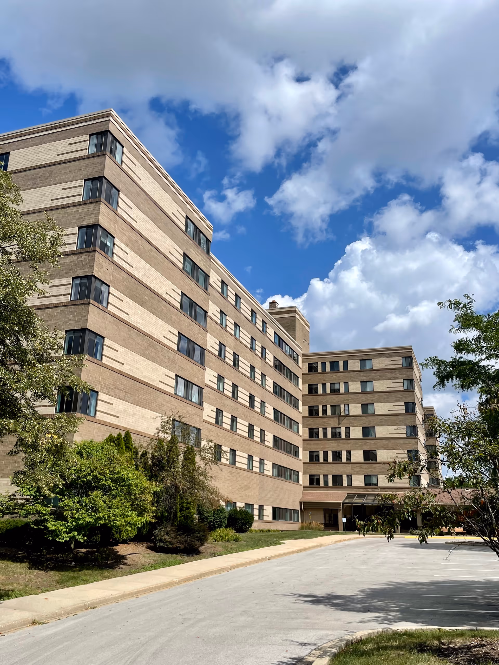 Exterior view of a multi-story senior living facility building under a partly cloudy blue sky. The building has a beige and light brown brick facade with many windows. There are trees and shrubs along the side of the building and a driveway leading to the entrance.