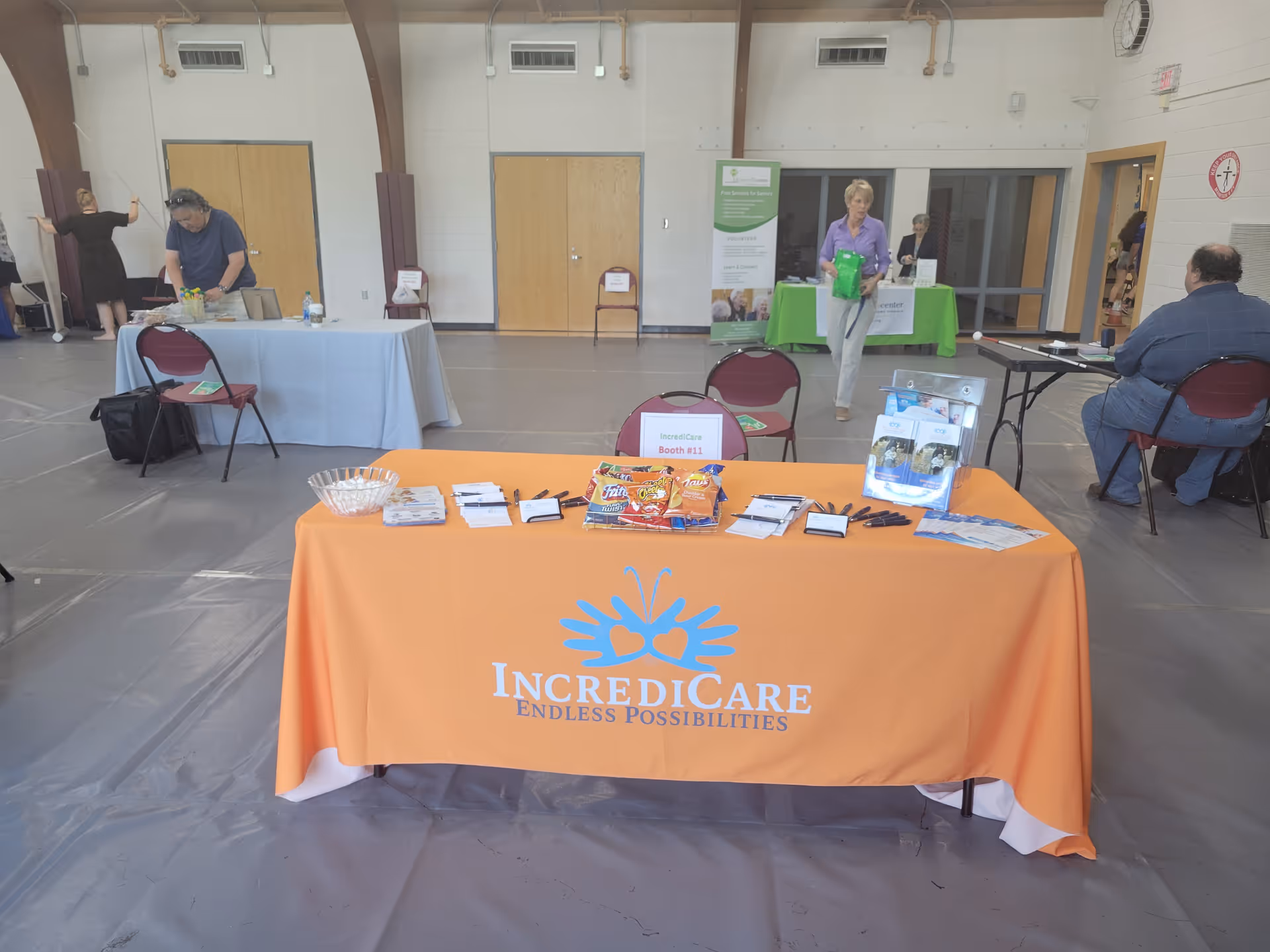 Indoor hall with orange IncrediCare table, brochures, and several people at booth displays.