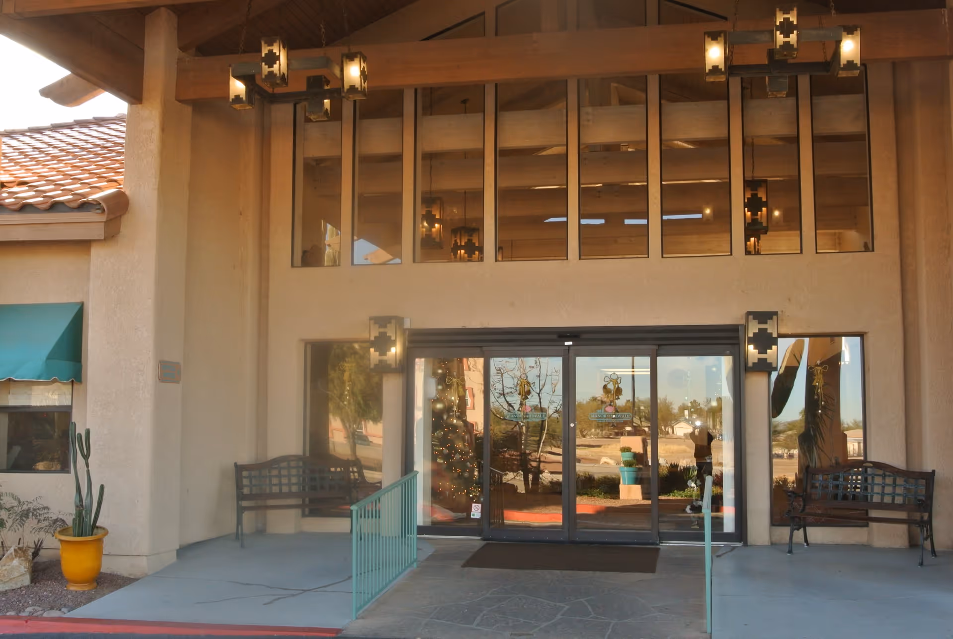 Entrance of a senior living facility with glass double doors, two benches on either side, a yellow pot with a cactus plant, and decorative hanging lights above. The building has a beige exterior and a tiled roof.