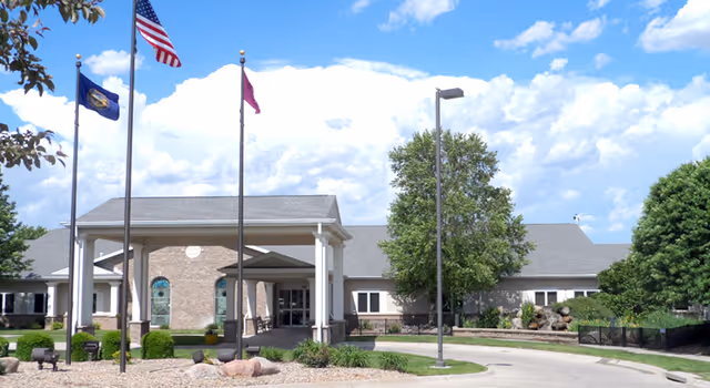 Front entrance of the South Haven building with a porte-cochère, flagpoles, landscaped grounds, and a circular driveway under a blue sky.