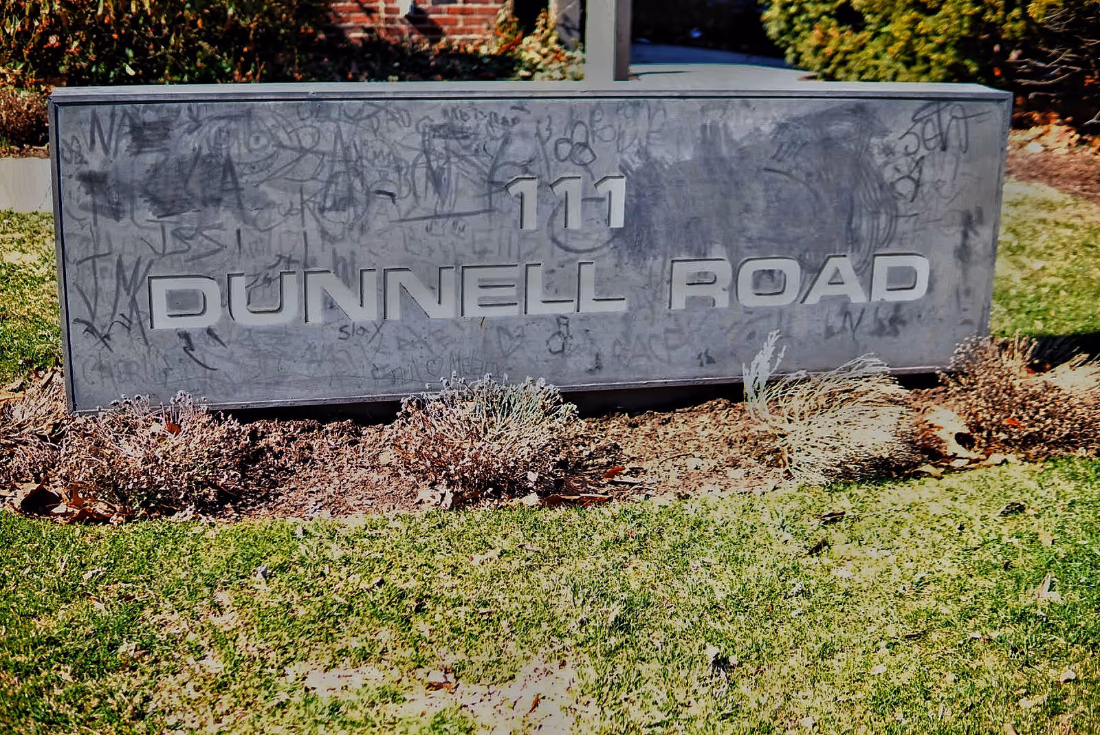 A large concrete street sign reading '111 Dunnell Road' with graffiti, set in a landscaped area.