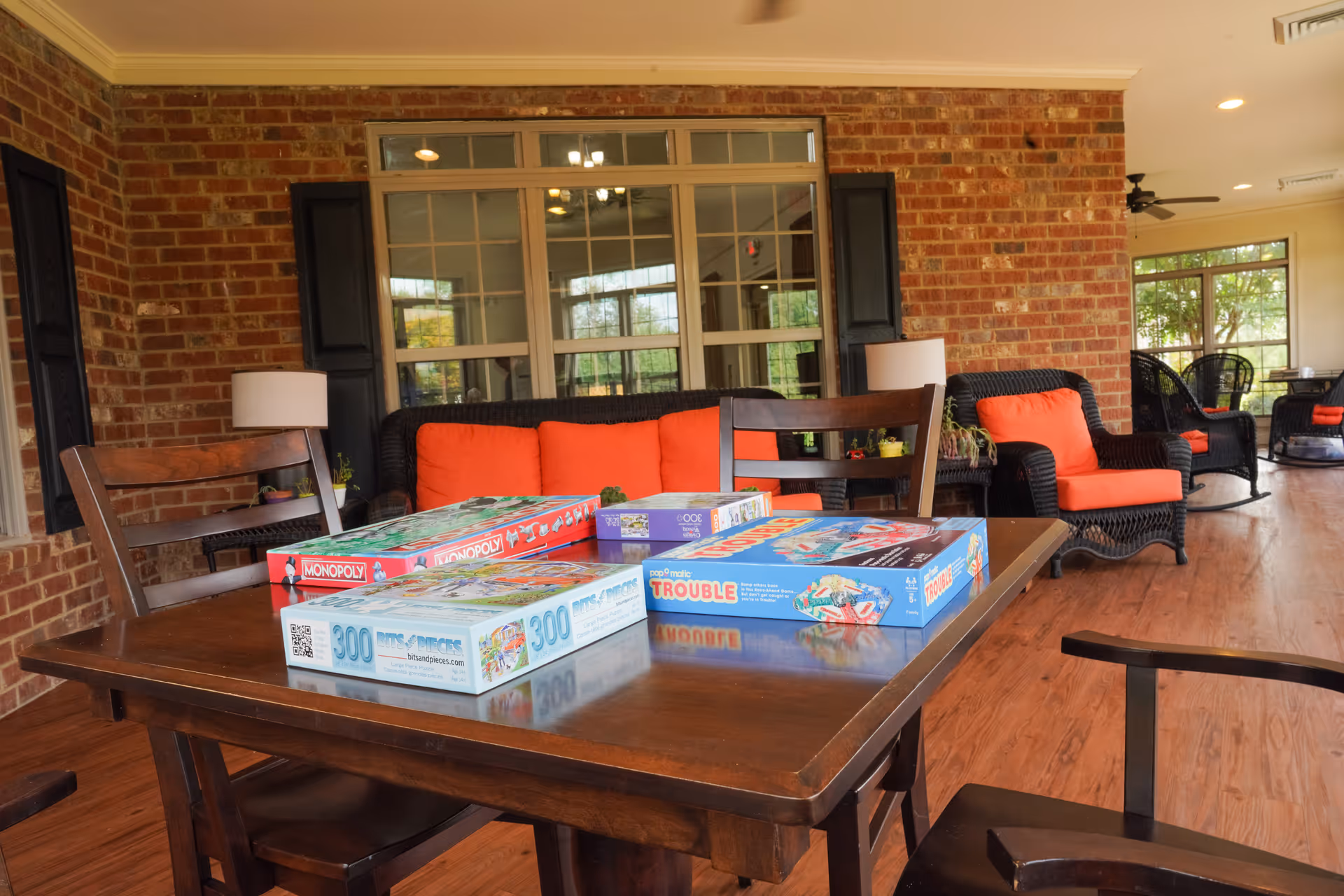 Indoor common room with a wooden table holding board games and wicker sofas with bright orange cushions against a brick wall and large windows.