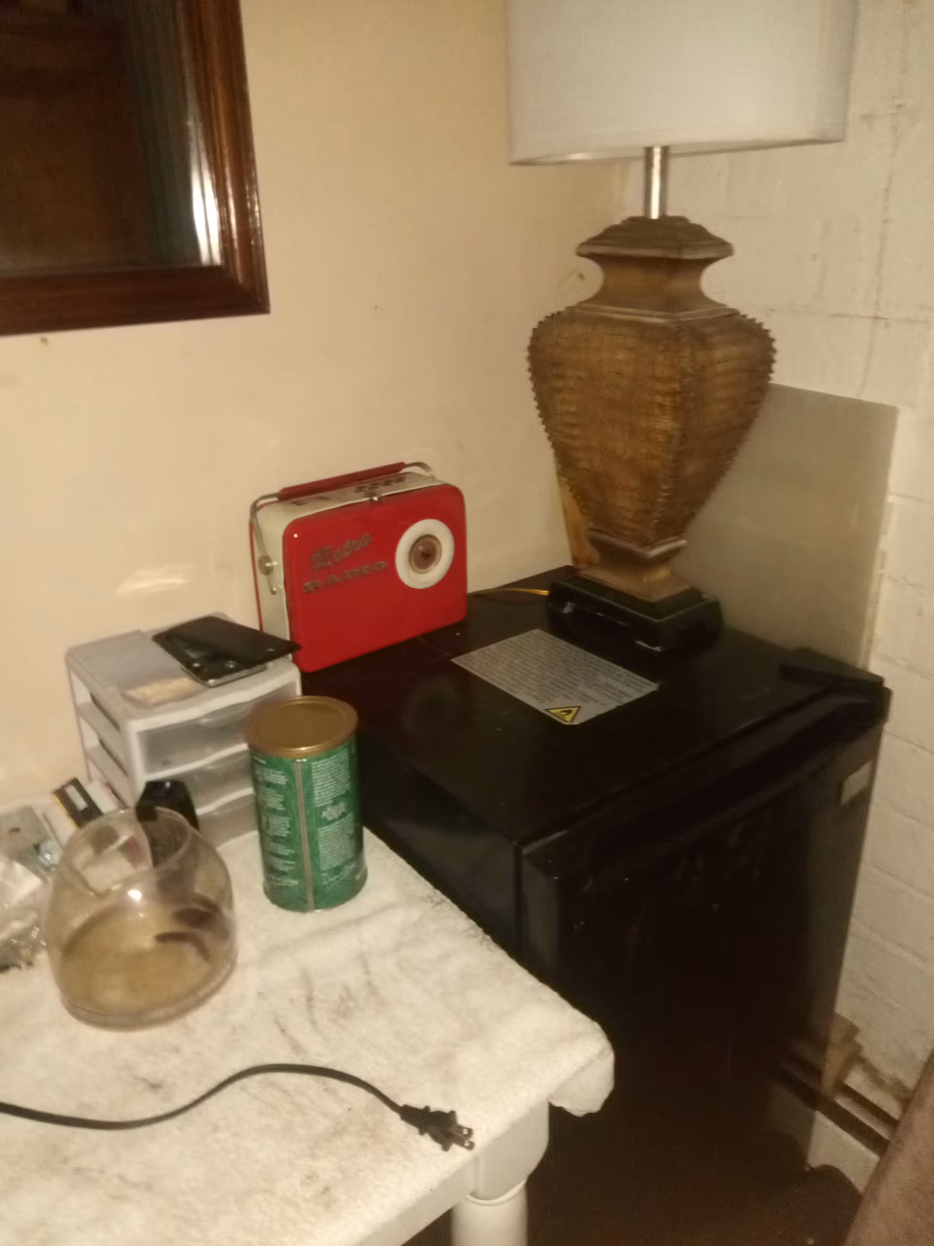 A cluttered small corner with a red vintage radio, lamp, mini-fridge, jars, and towels.