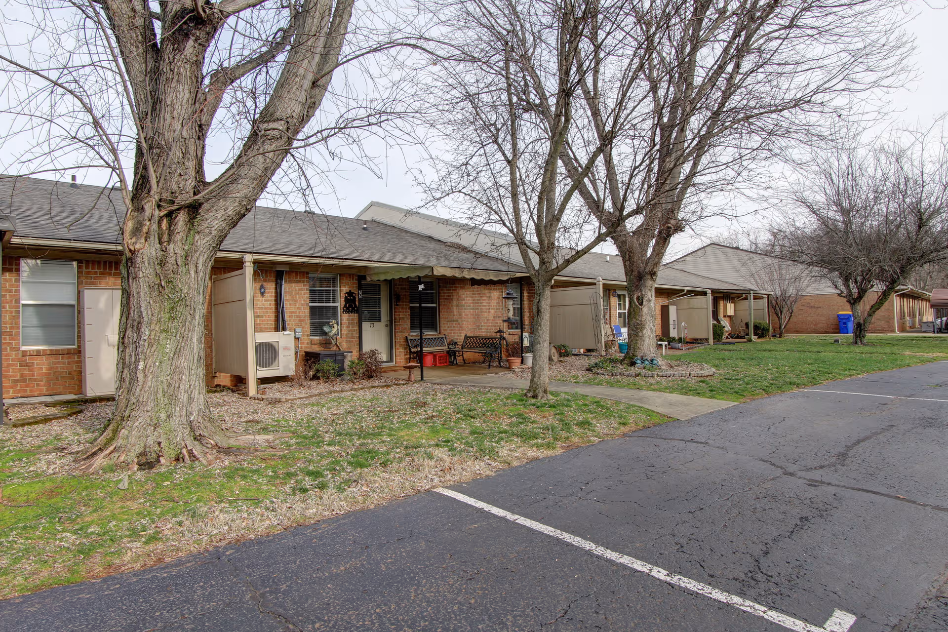 Row of brick senior living cottages with porches, trees, and a paved parking lot.