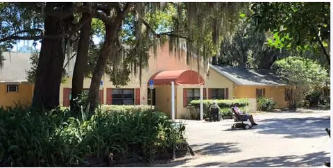 Front entrance of a single-story yellow senior living building with a red awning, trees, landscaping, and a person on a mobility scooter outside.