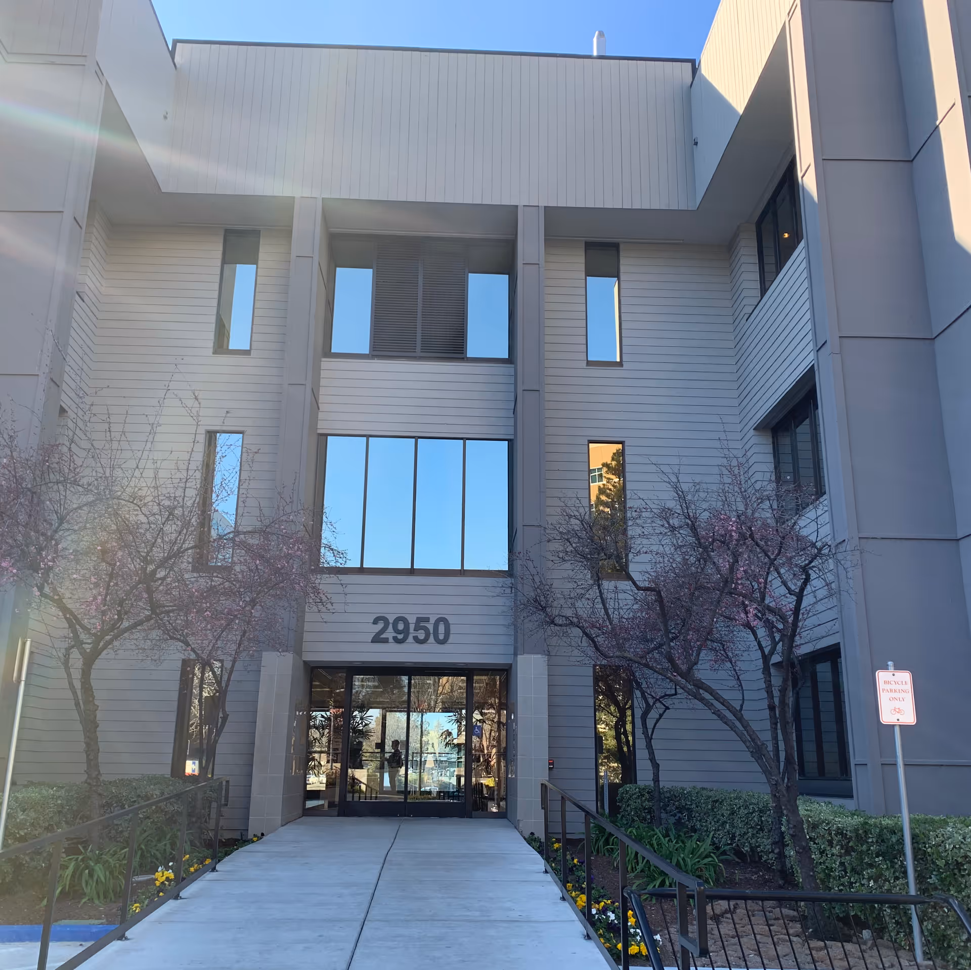 Modern three-story facility entrance with glass doors, numbered 2950, surrounded by blooming trees.