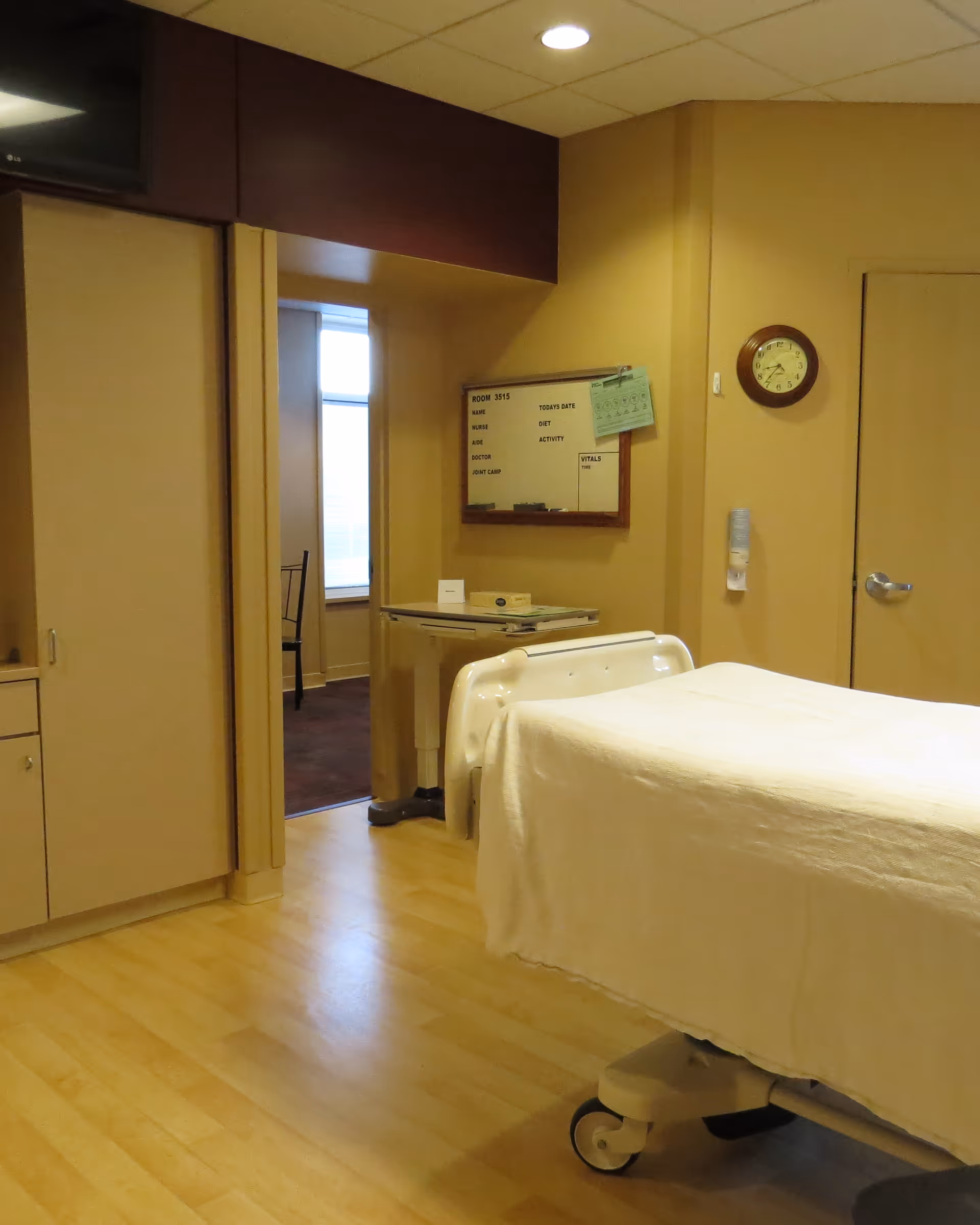 A hospital bed in a beige interior patient room with white linens and a wall clock.