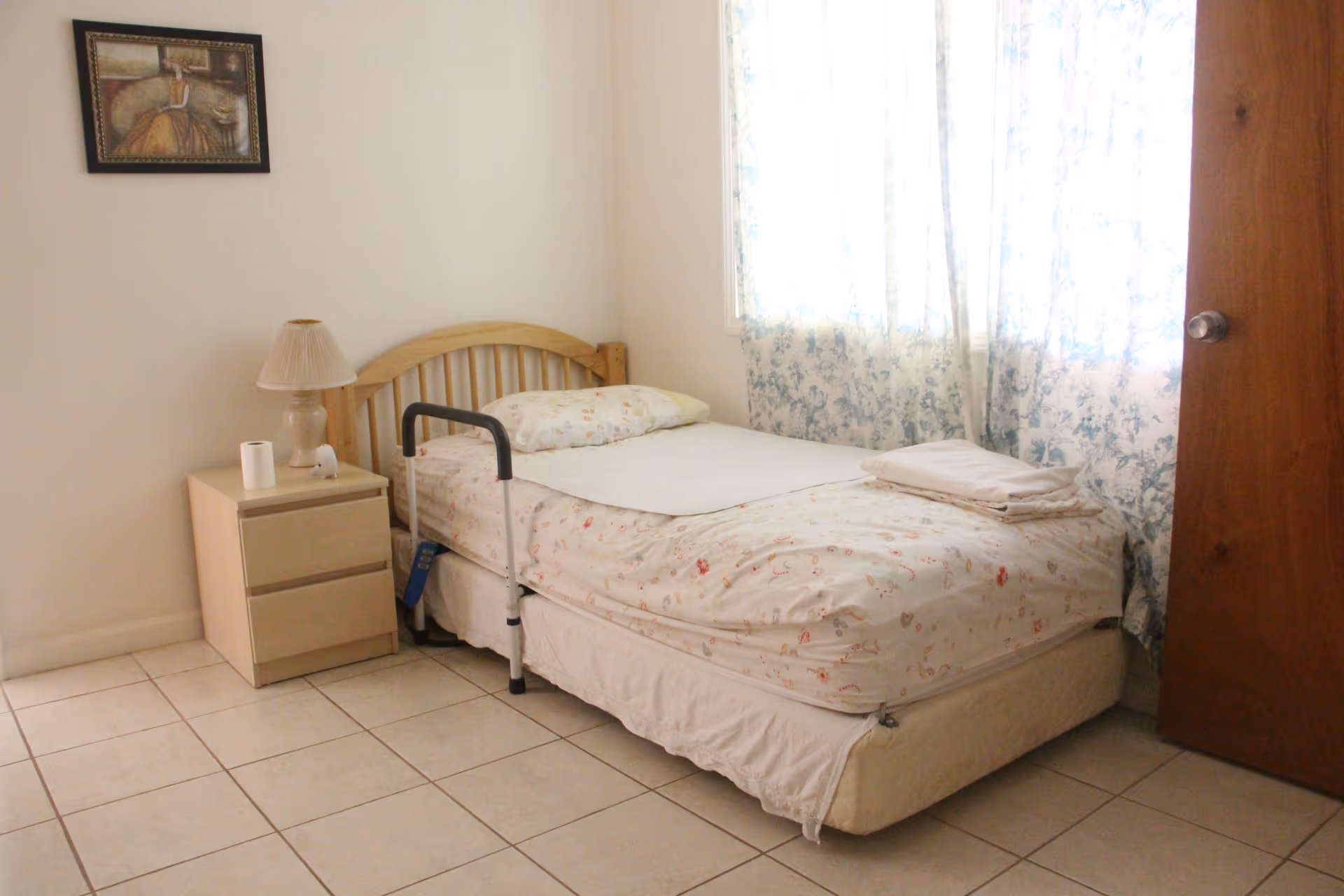 A simple bedroom with a bed, nightstand, lamp, and floral curtains in a senior living facility.