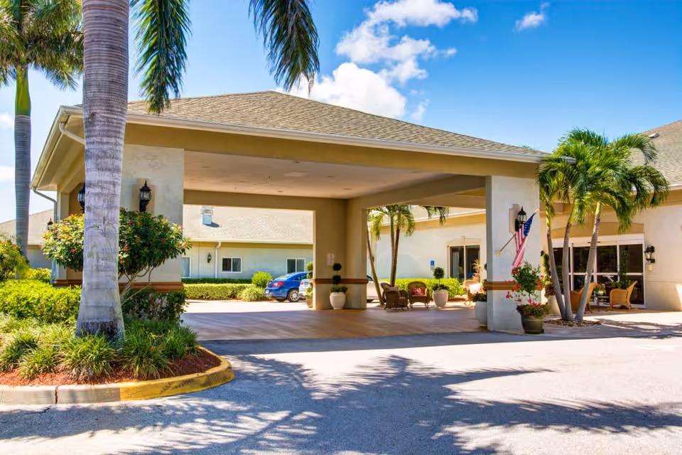 Entrance area of a senior living facility with a covered drop-off zone, palm trees, potted plants, outdoor seating, and an American flag under a clear blue sky.