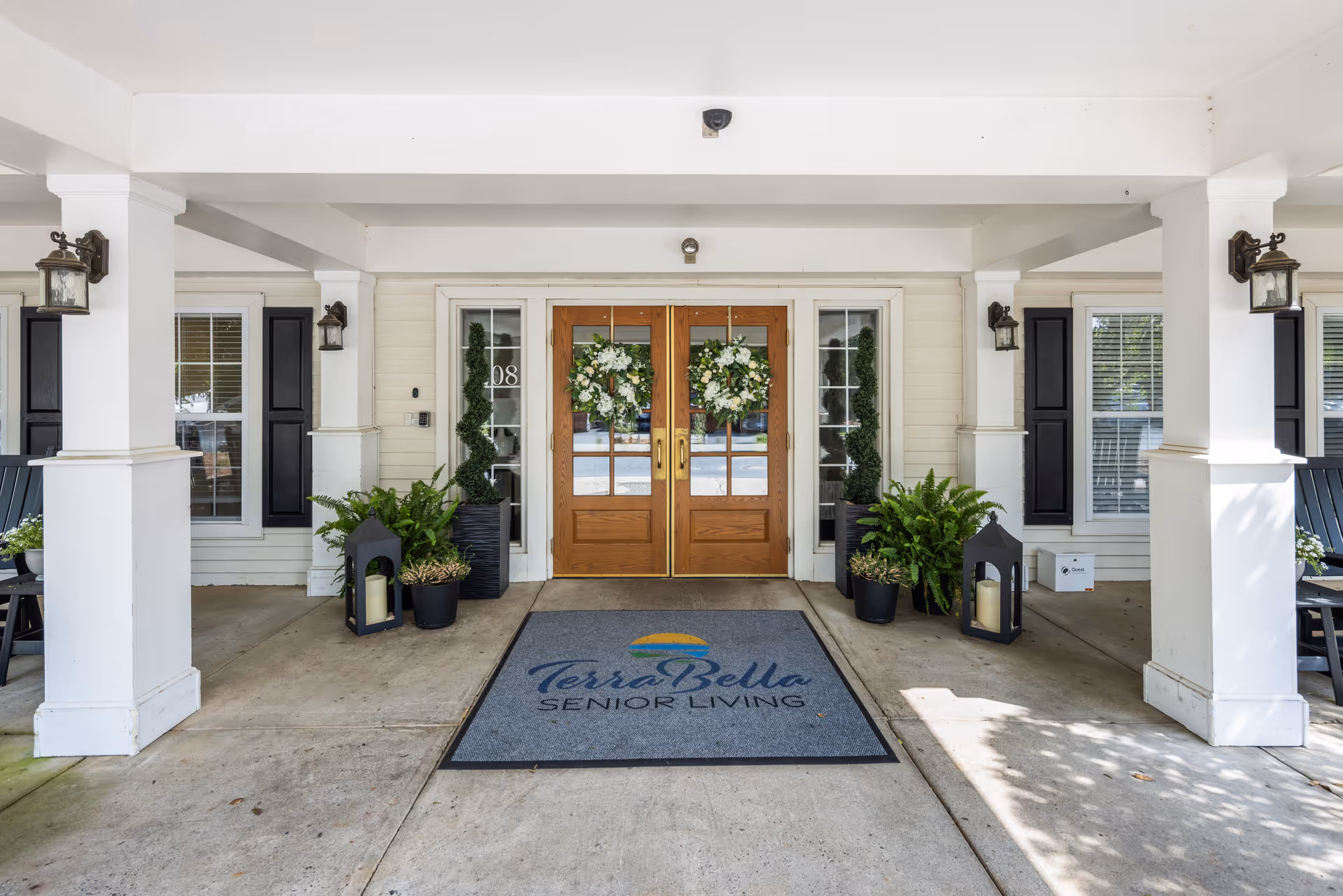 Entrance to TerraBella Greensboro senior living facility with double wooden doors decorated with floral wreaths, flanked by tall plants and lanterns. A welcome mat with the TerraBella Senior Living logo is placed in front of the doors under a covered porch supported by white columns.