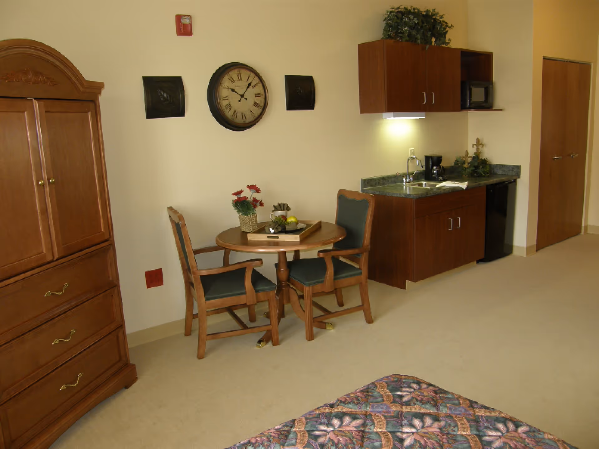 Studio apartment interior showing a small dining table with two chairs, a kitchenette, armoire, and part of a bed.