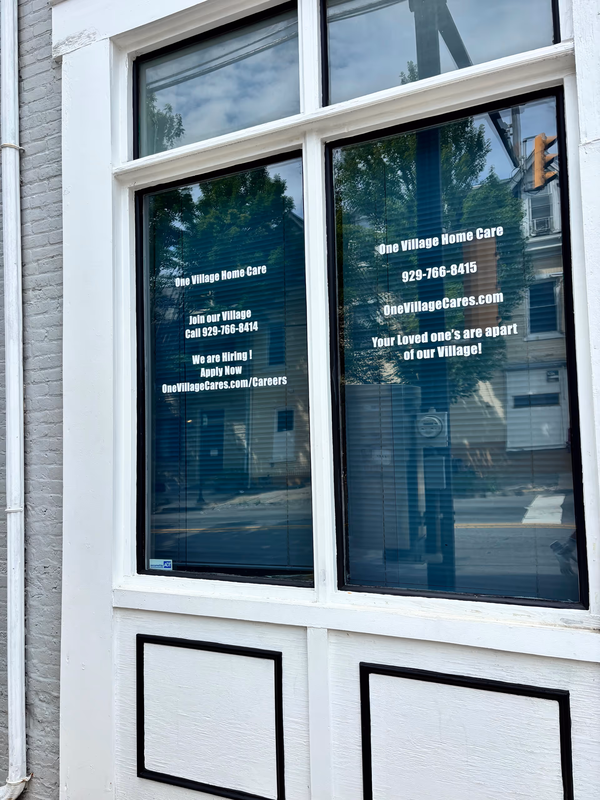 White-framed storefront window displays One Village Home Care signage and contact information.