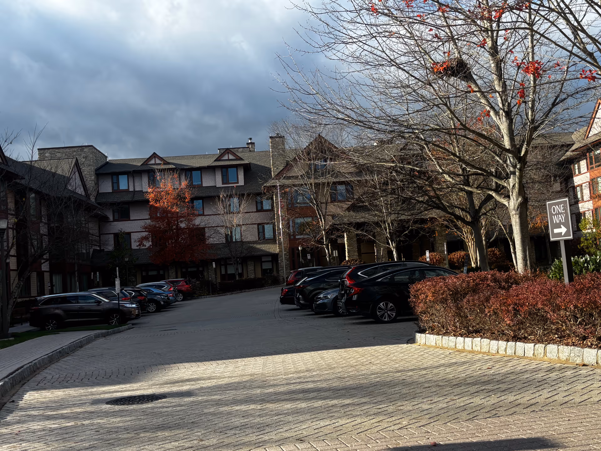Front exterior of a multi-story senior living building with parked cars, bare trees, and a 'One Way' sign.