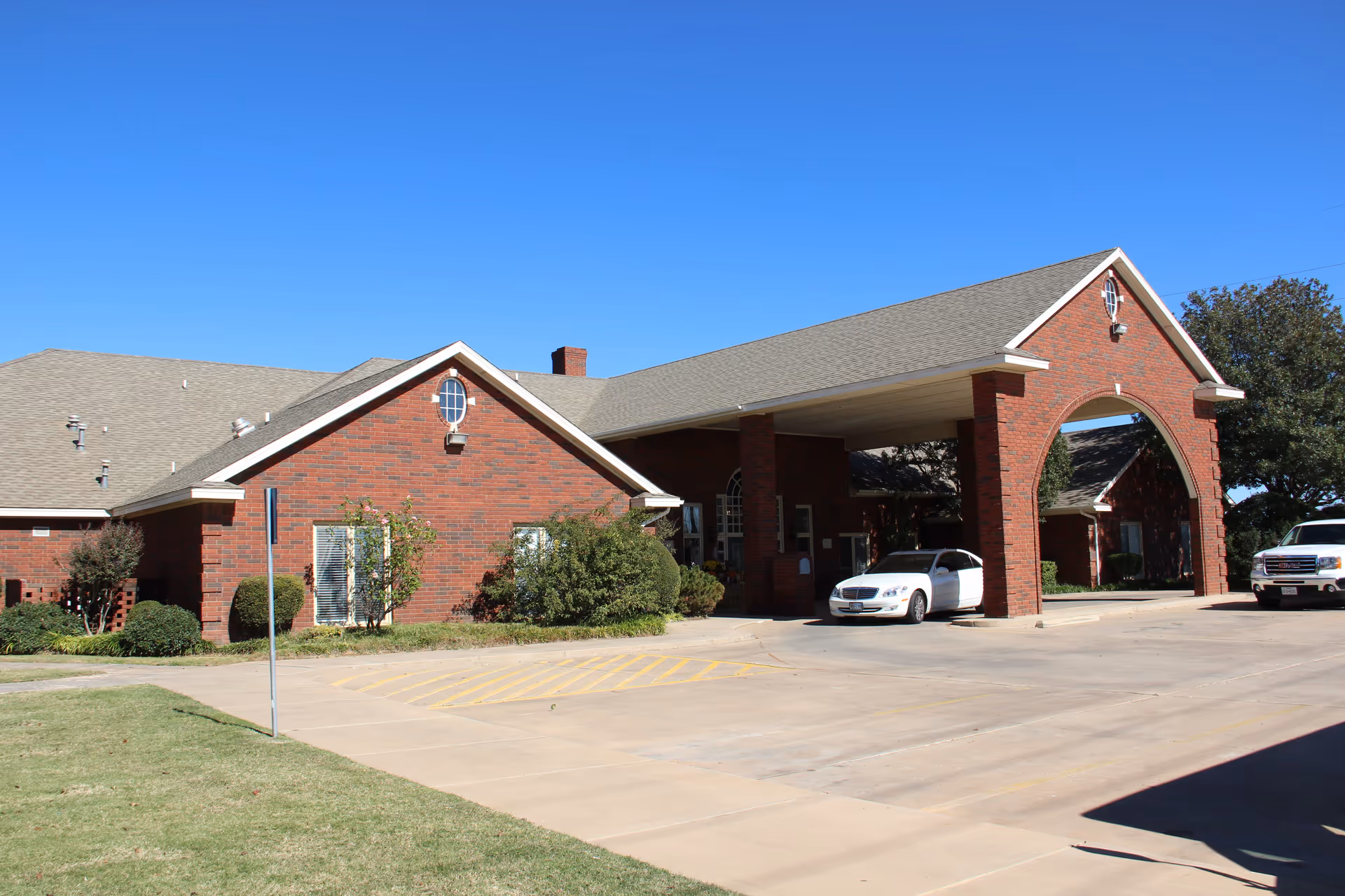 Brick senior living facility entrance with a covered drive, arched gateway, and parked cars.
