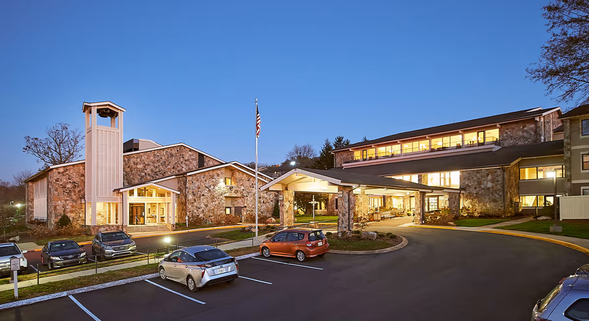 Exterior view of Ascension Living Alexian Village - Tennessee assisted living facility at dusk, showing a stone building with a covered entrance, several parked cars, an American flag on a flagpole, and illuminated windows.
