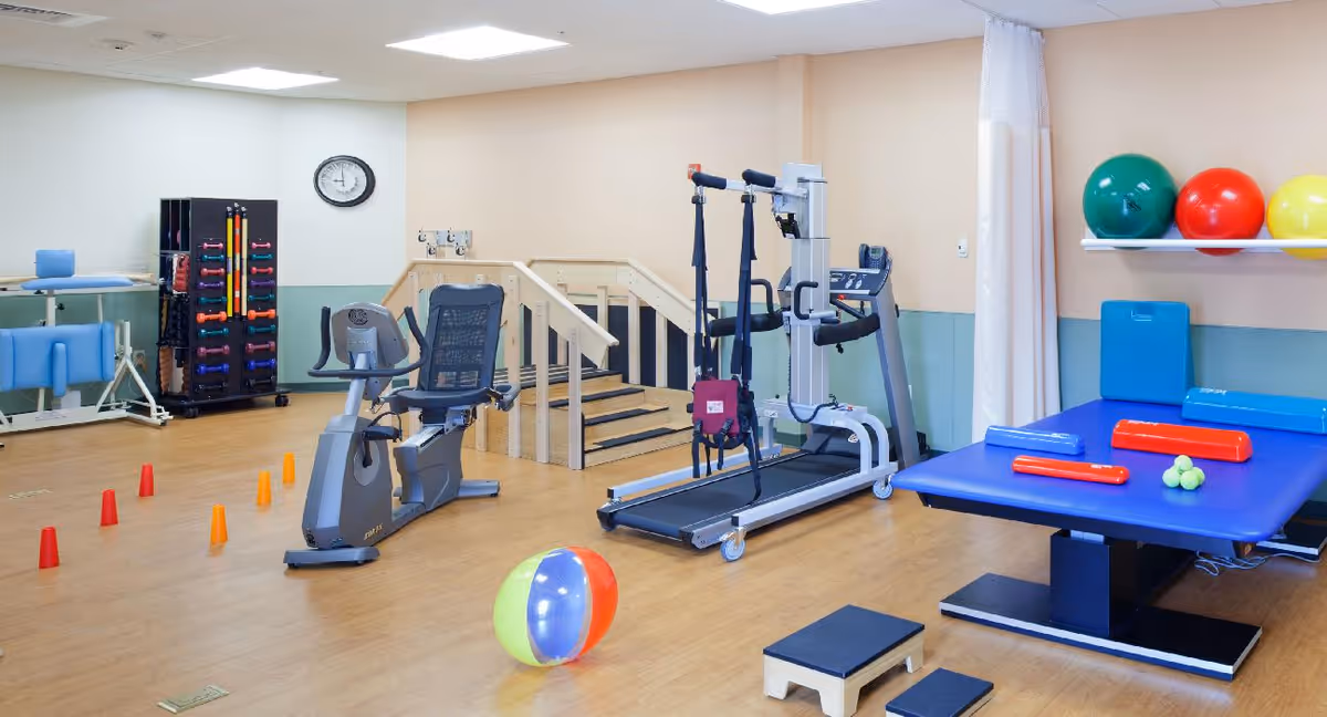A rehabilitation therapy room with exercise equipment including a recumbent bike, treadmill with harness support, therapy tables, colorful therapy balls, small cones arranged on the floor, and various therapy tools on shelves. The room has light-colored walls and wooden flooring.
