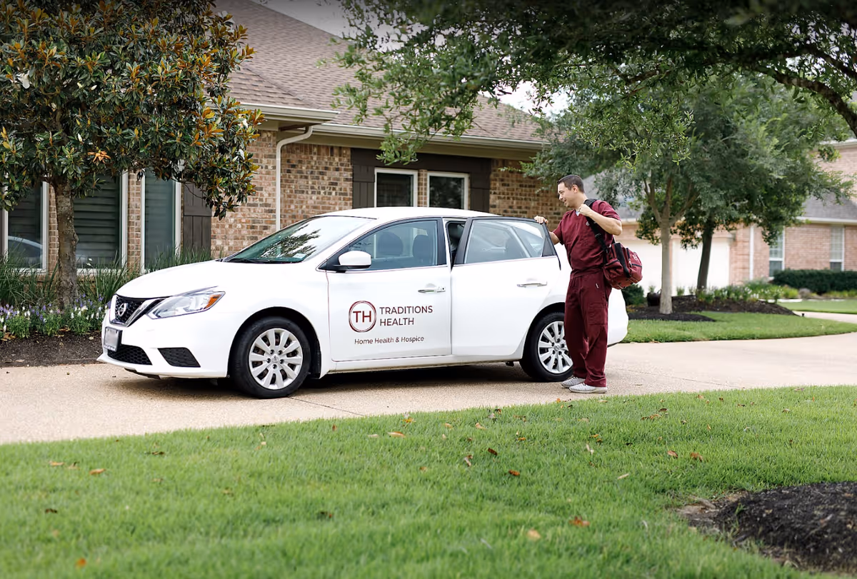 White car with Traditions Health logo parked outside brick senior care facility, staff member nearby.