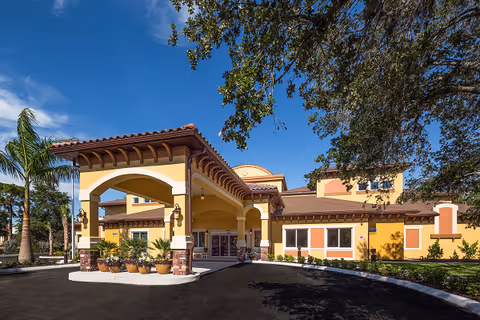 Exterior view of Aravilla Sarasota senior living facility showing a large covered entrance with arches, yellow and brown walls, and surrounding landscaping including palm trees and other greenery under a blue sky.