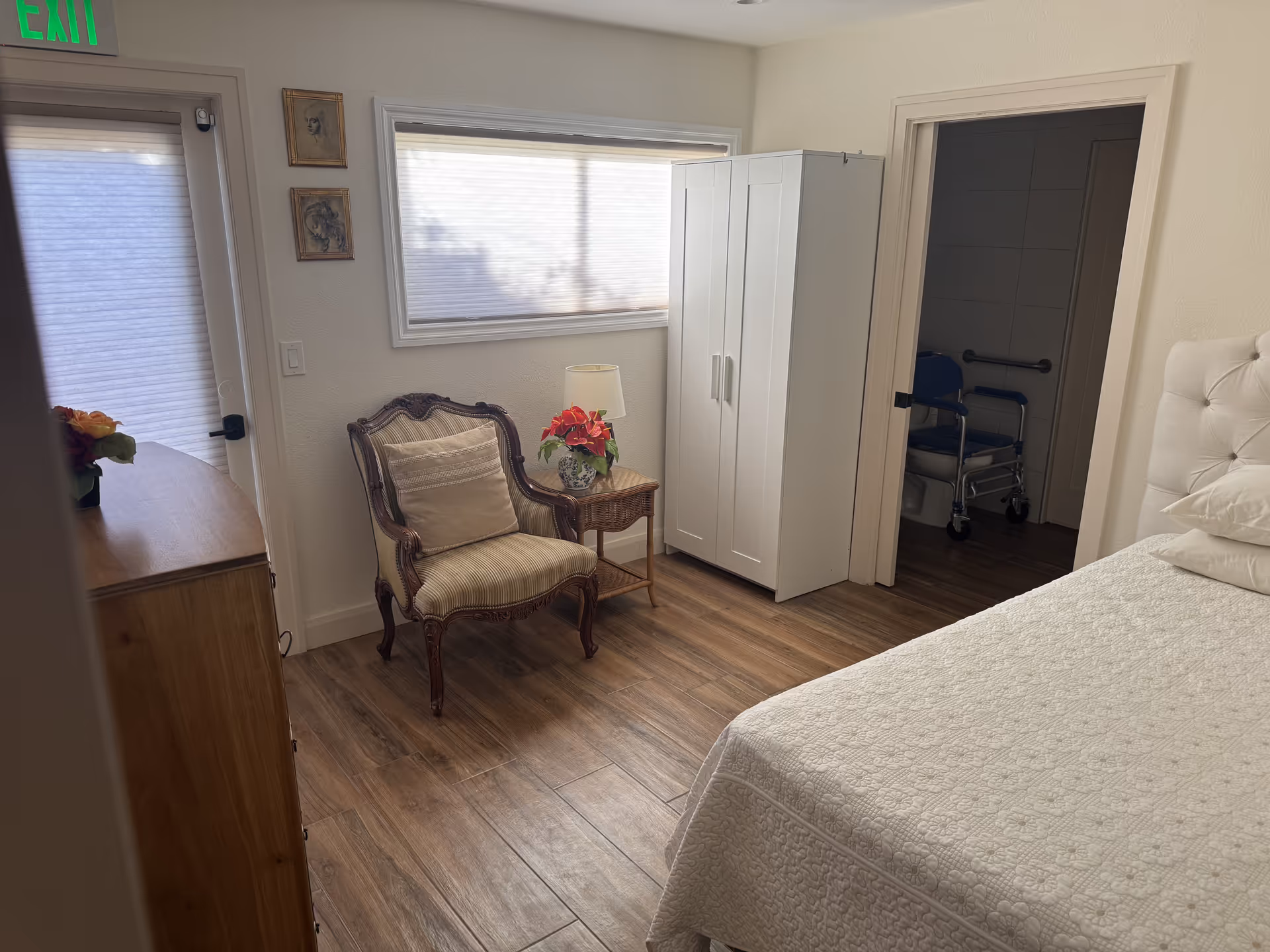 Bedroom in a senior facility with bed, chair, wardrobe, and walker visible near doorway.