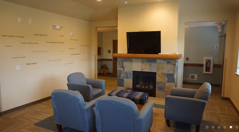 A cozy living room area in an assisted living facility featuring four blue upholstered armchairs arranged around a striped ottoman in front of a stone-tiled fireplace with a flat-screen TV mounted above it. The walls are beige with various quotes displayed on one side, and there is a hallway visible in the background.