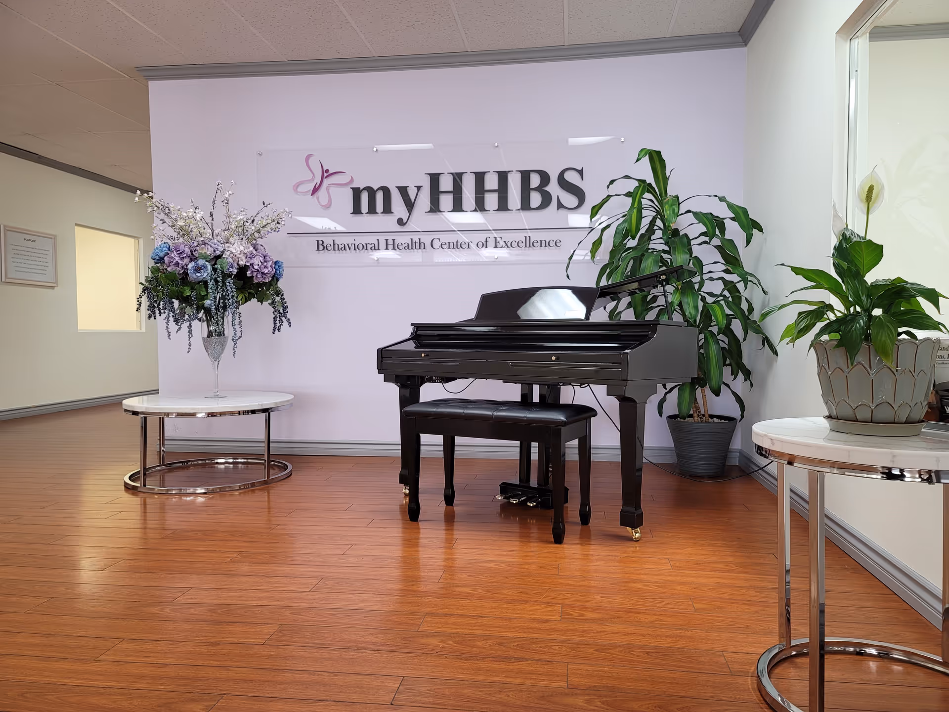 A black grand piano sits in a lobby with plants and a MyHHBS sign.