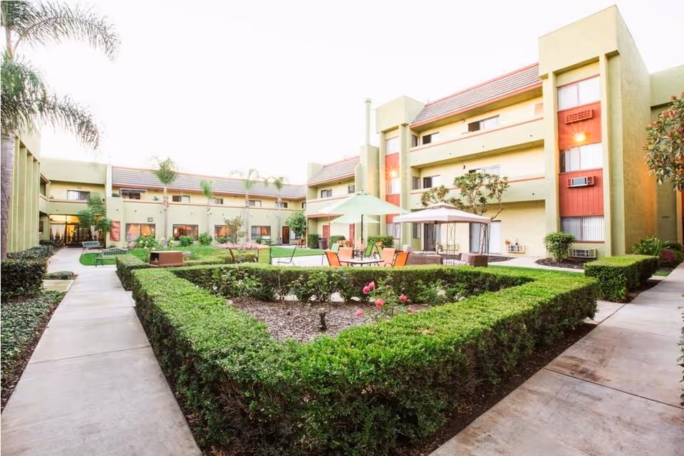 Outdoor courtyard area of Vista Del Mar Senior Living featuring trimmed hedges, flower beds, patio tables with umbrellas, chairs, and a gazebo. The courtyard is surrounded by a three-story building with windows and balconies.