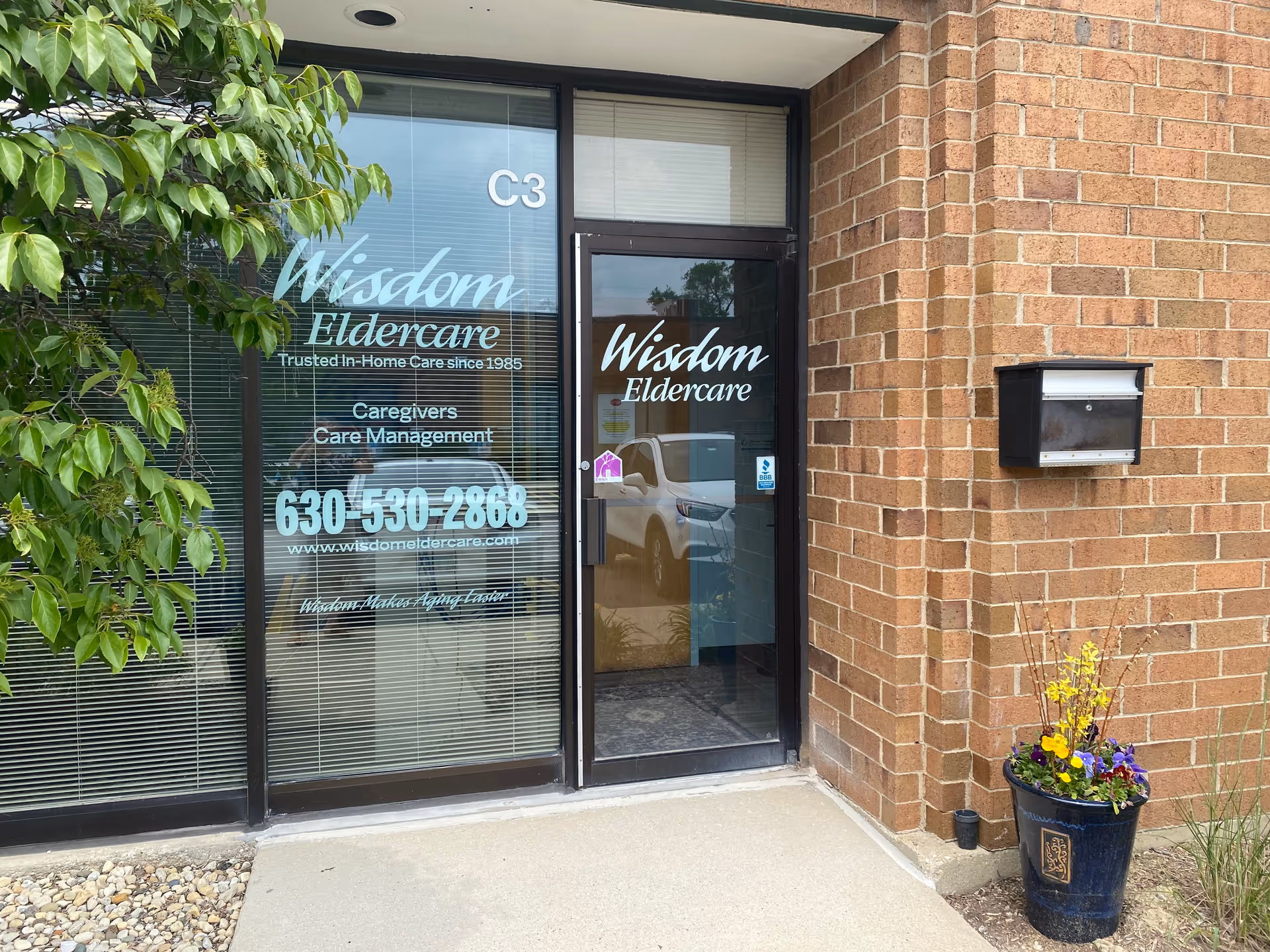 Brick building entrance with Wisdom Eldercare glass door and potted flowers outside.