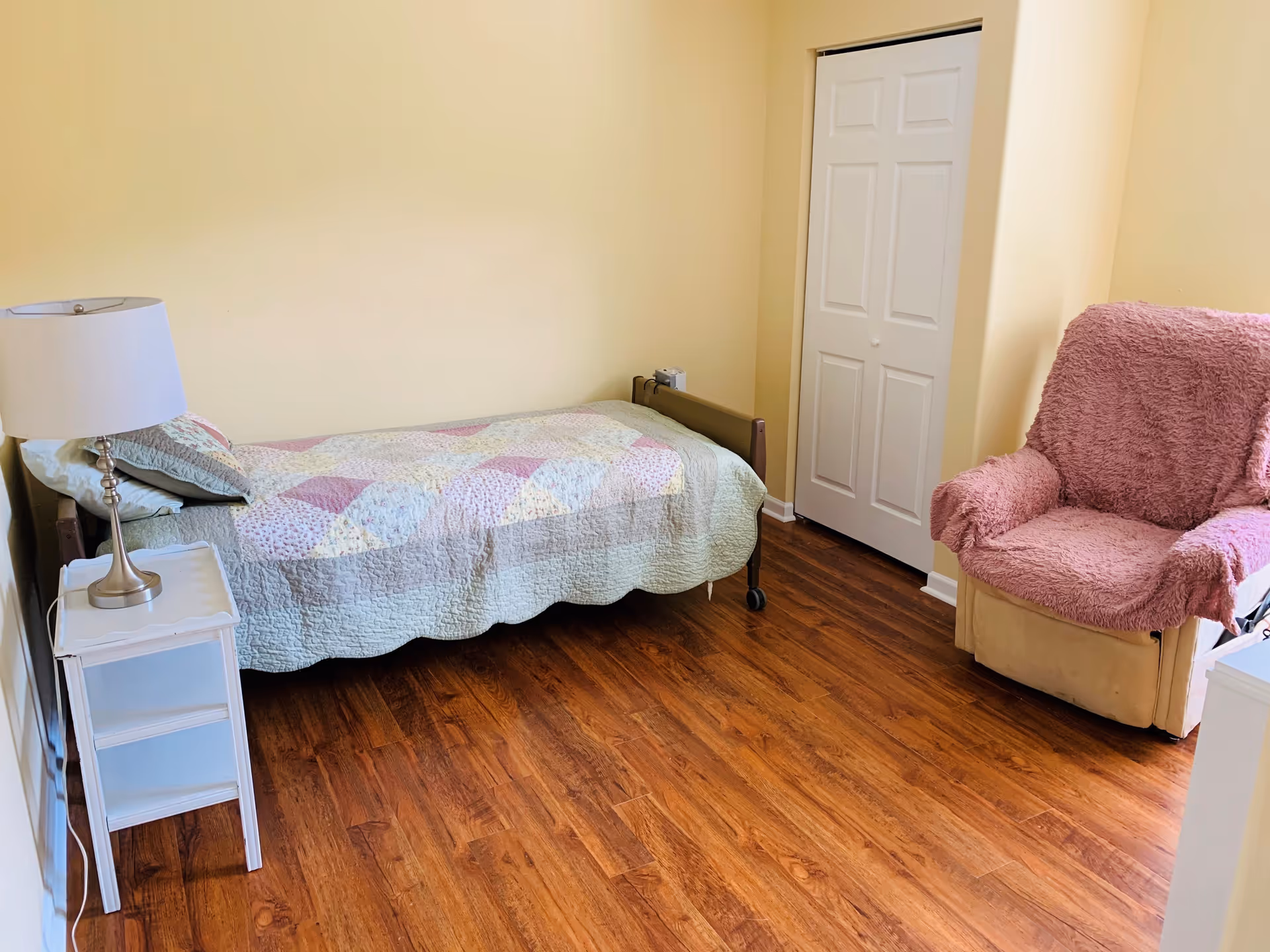 A small bedroom with a single bed covered with a pastel patchwork quilt, a white nightstand with a lamp, a pink cushioned armchair with a fuzzy cover, and a closed white closet door. The room has light yellow walls and wooden flooring.