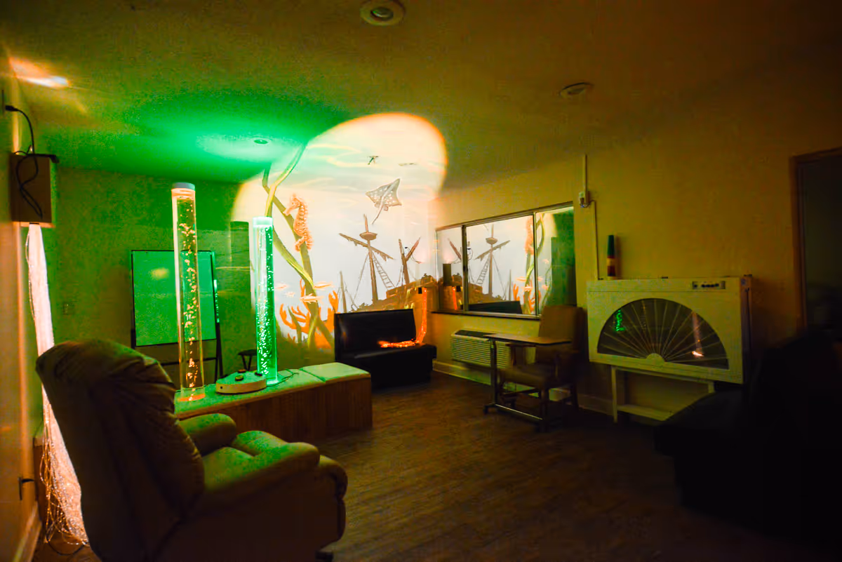 A dimly lit room with a cozy recliner chair, a black cushioned bench, and a small table with a chair. The wall features a colorful underwater mural with sea creatures and a shipwreck. Two tall bubble tubes with green lighting add a calming ambiance to the space.