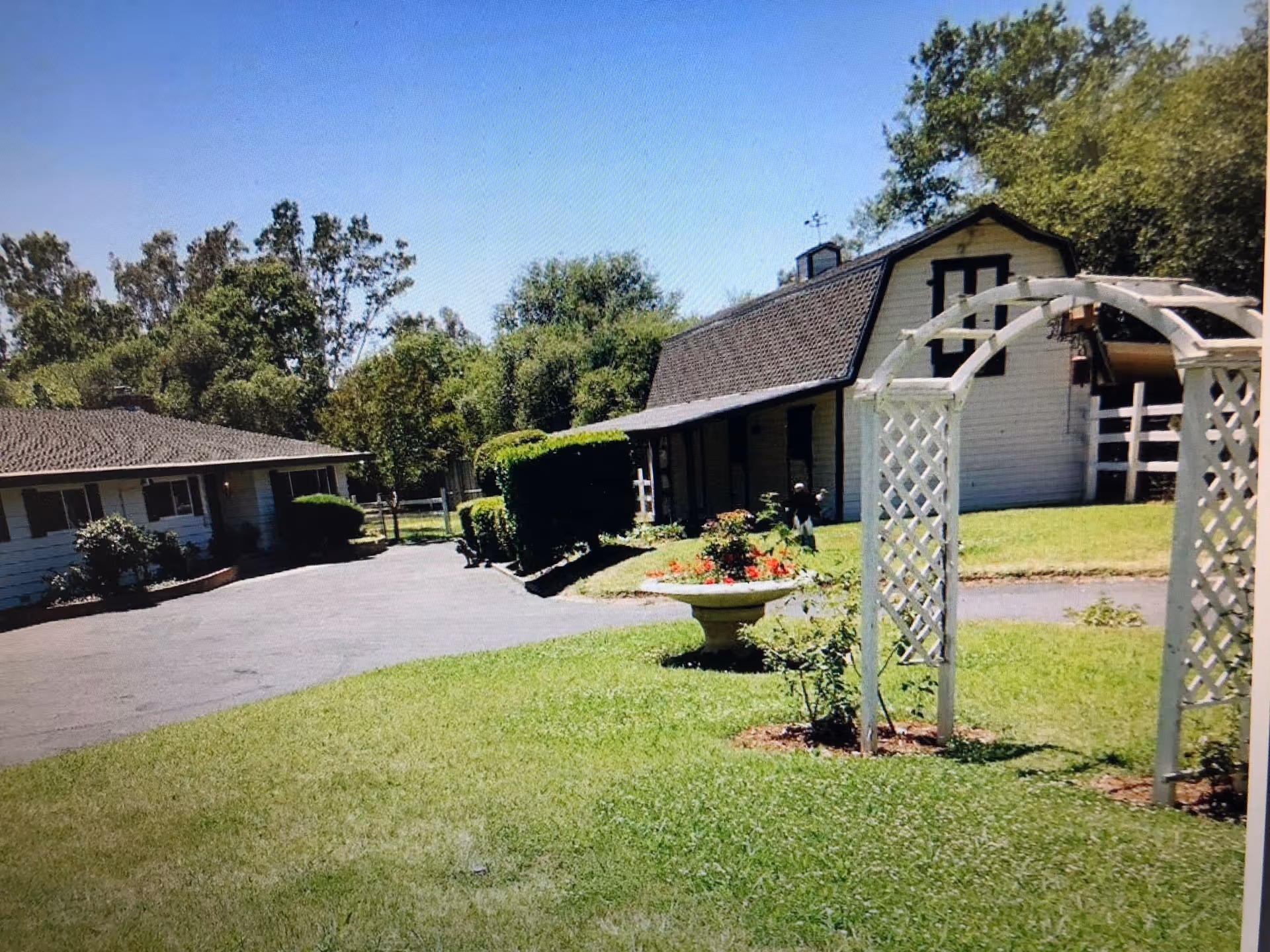 Front view of a ranch-style property with a driveway, manicured lawn, trellis arch, planter, and two white buildings.
