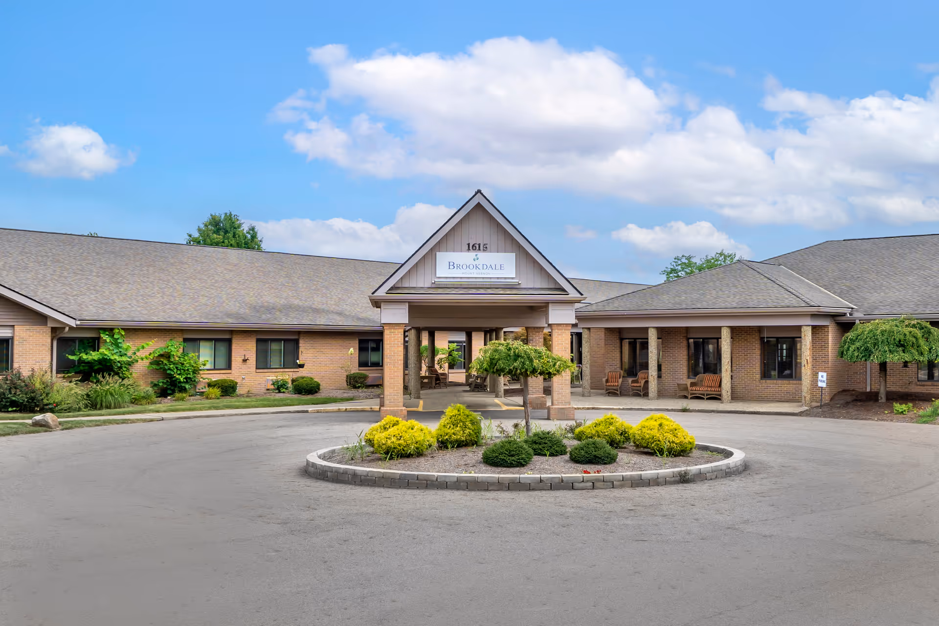 Front exterior view of Brookdale Mount Vernon facility with a circular driveway and landscaped roundabout featuring shrubs and small trees under a partly cloudy blue sky.