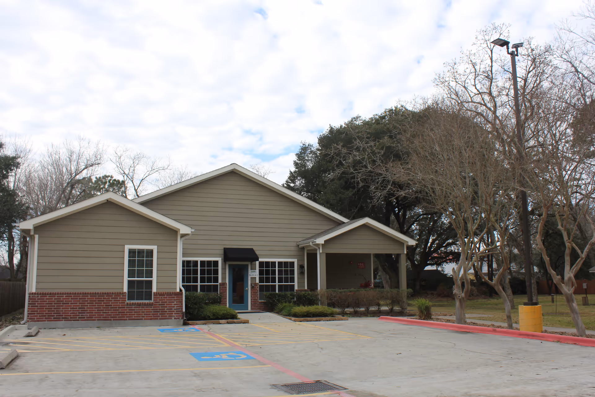 Exterior view of a single-story building with beige siding and red brick accents, featuring a small covered entrance and surrounded by leafless trees and a parking lot with handicap parking spaces.