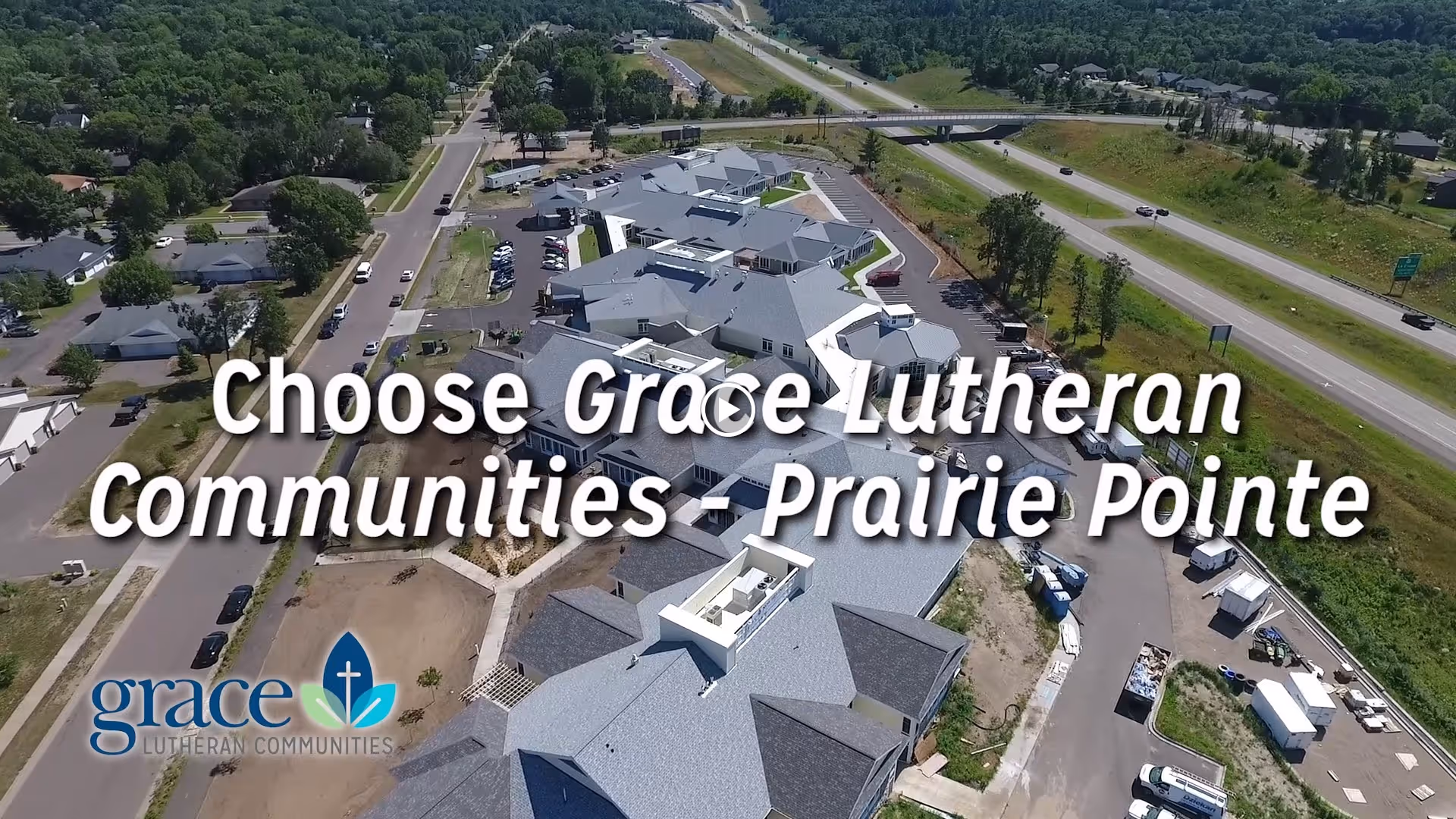 Aerial view of Grace Lutheran Communities - Prairie Pointe Assisted Living & Memory Care facility showing multiple connected buildings with gray roofs, surrounding roads, parked cars, and greenery in the background.