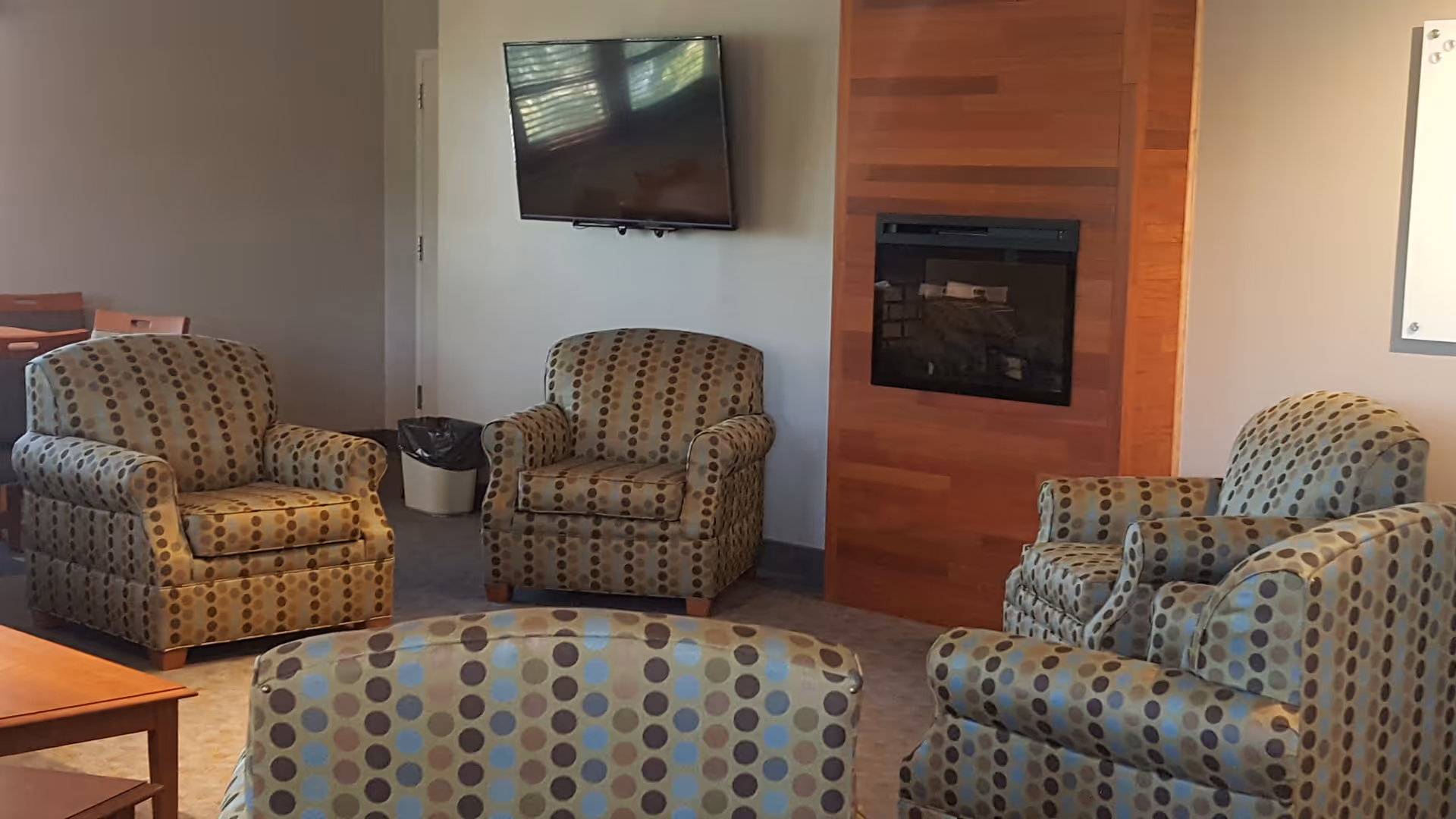 Cozy senior living lounge with patterned armchairs, a wall-mounted TV, and a wood fireplace.