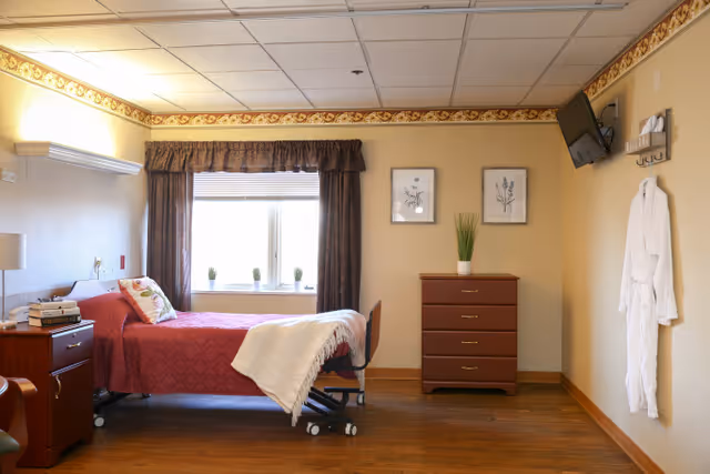 A furnished senior living bedroom with a bed, dresser, window, and robe hanging.