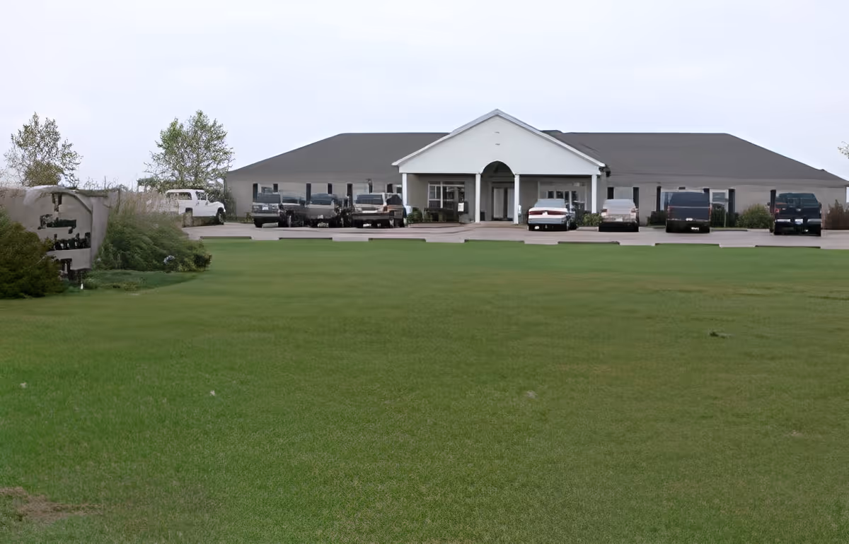 Single-story residential care building with a large grassy lawn and parked cars in front of the entrance.