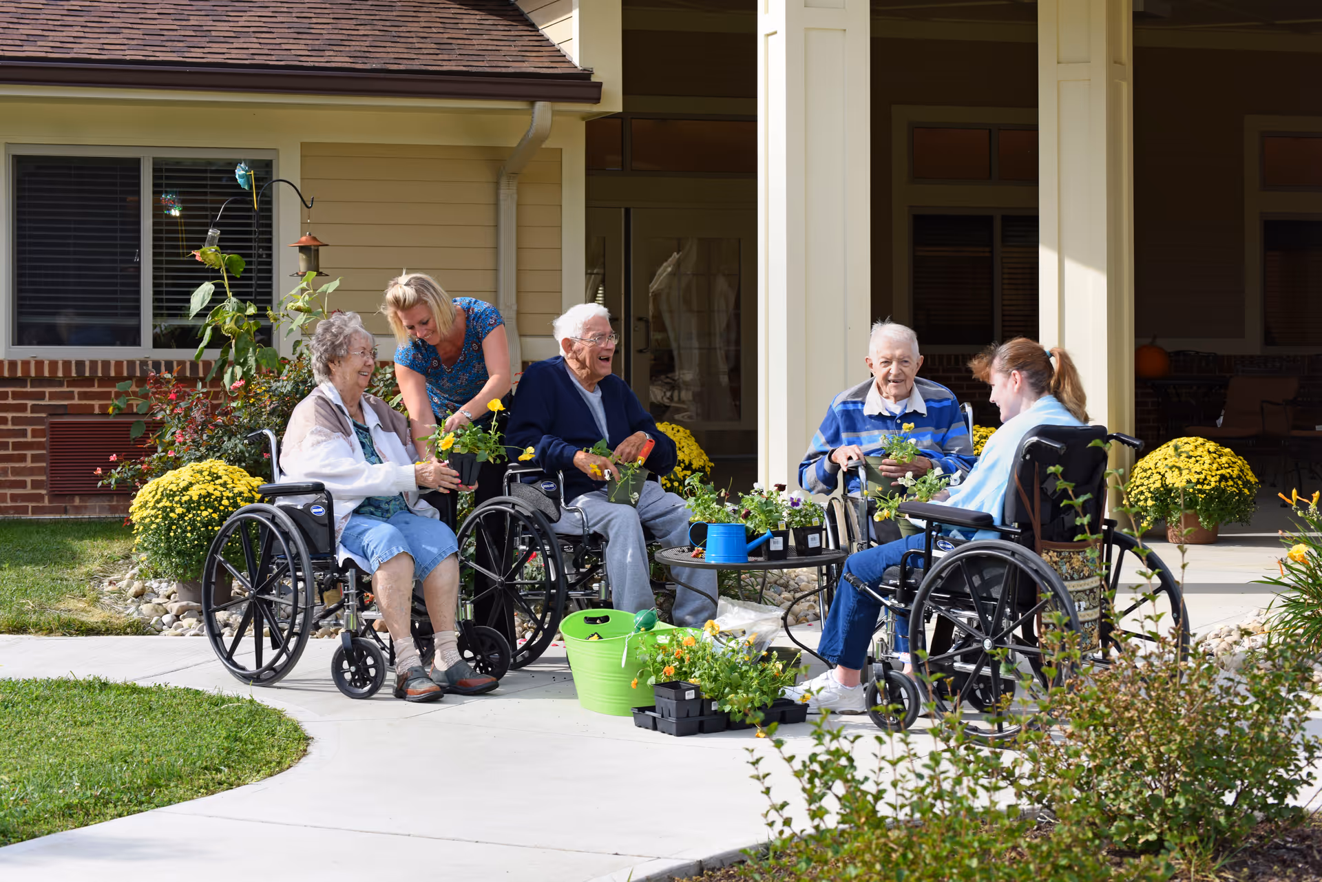 Four elderly individuals in wheelchairs and a caregiver are outside a nursing facility, engaging in a gardening activity with potted plants and flowers on a sunny day.