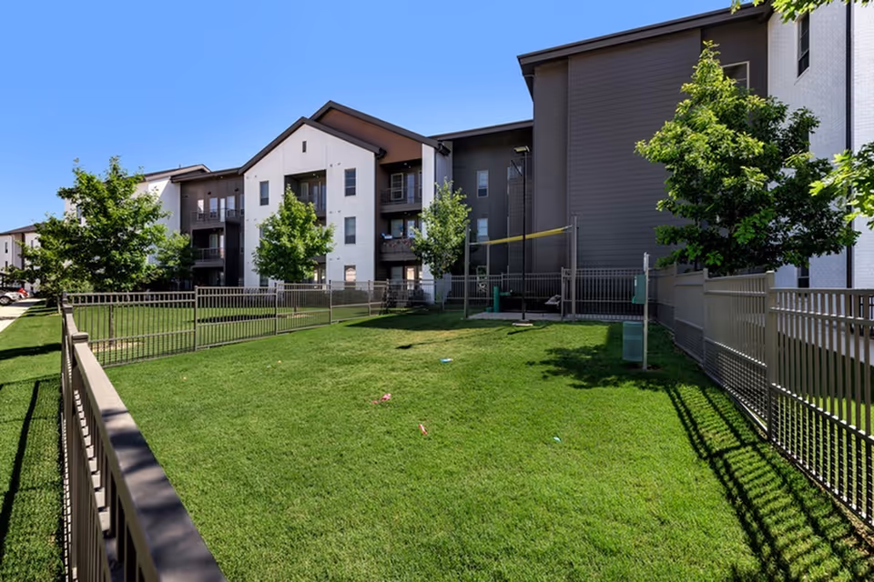 Fenced grassy courtyard in front of a multi-story senior living building with balconies and trees under a clear blue sky.