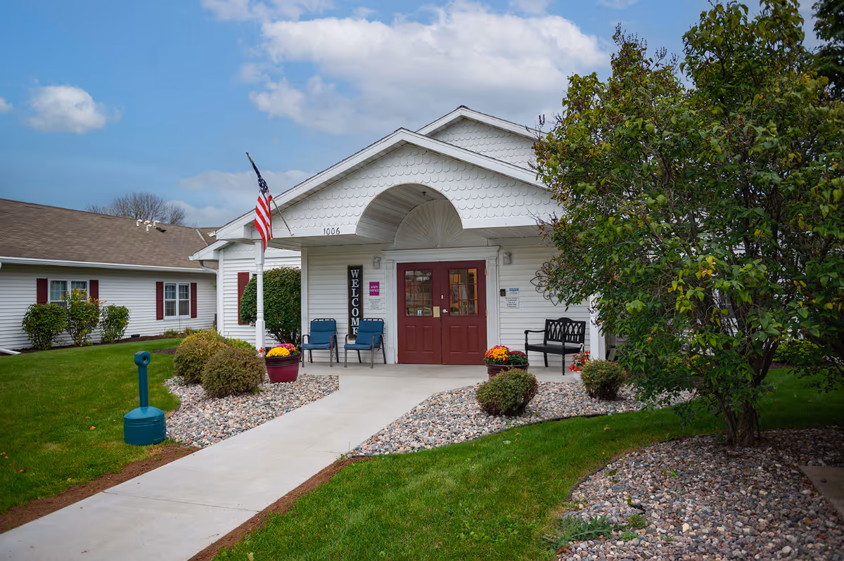 Exterior view of a white senior living facility with red doors, welcome sign, and landscaping.