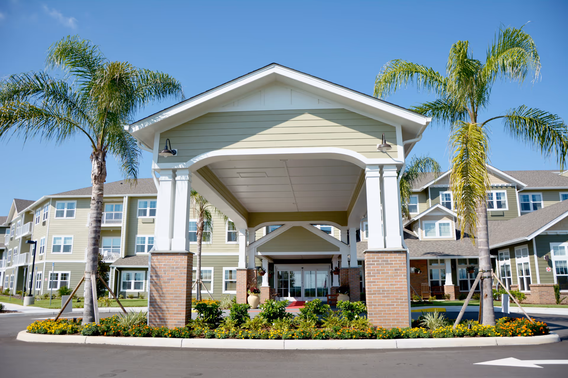 Front exterior view of Orchard Heights Gracious Retirement Living building with a covered entrance, palm trees, and landscaped flower beds under a clear blue sky.