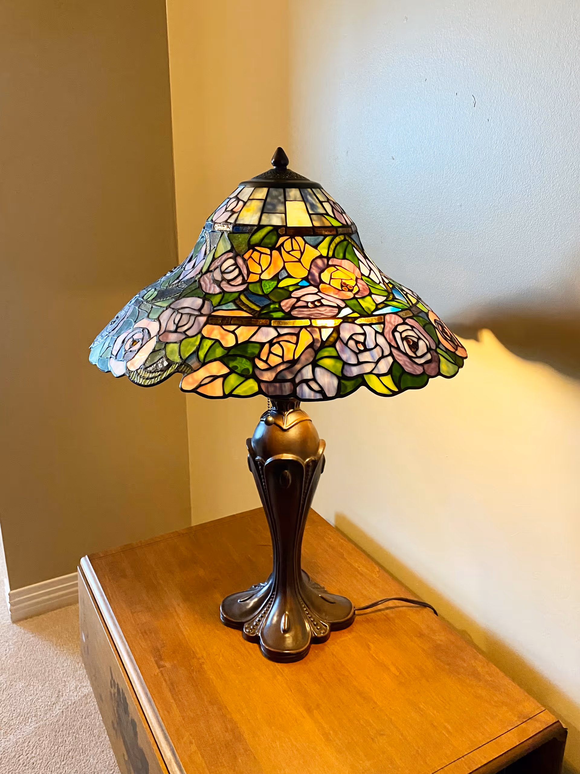 A decorative table lamp with a stained glass shade featuring a floral pattern in colors including pink, yellow, green, and purple, sitting on a wooden surface against a beige wall.
